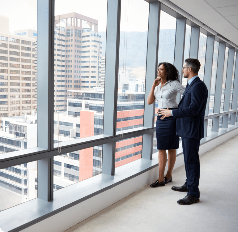 Two people standing at a big glass wall looking out at a city